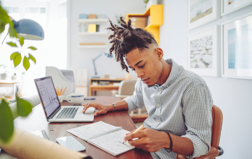 A young man working at a desk with a notepad and laptop.