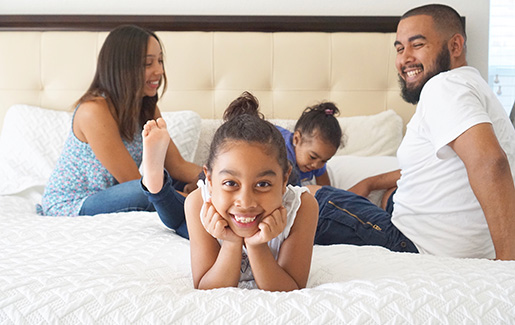 A mom, dad and two little girls smile while playing on a bed