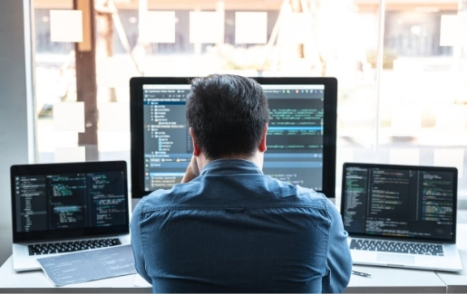 Male in blue shirt working in front of the window during the day with three computers