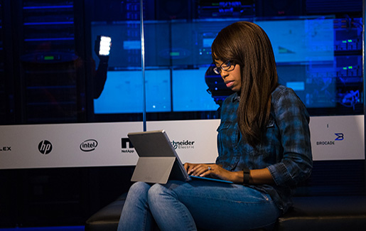 A female cybersecurity pro works on a laptop near the server room. Photo by WOCinTechChat.com.
